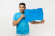 © khosrork - Joyful unshaven man wearing blue T- shirt standing holding speech or think blue frame, empty communication space, looking at camera with smile. Indoor studio shot isolated on gray background.