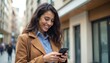 © Viktor - Smiling brunette woman uses smartphone outdoors on city street. She holds phone in hands, looking down at screen. Person wears coat and collared shirt. Background shows blurred urban buildings.