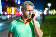 © Volodymyr - Man talk on phone in night street. Tourist calling on phone in night city. Hispanic guy using phone. Mature man talking on phone during travel. Urban tourist conversation on smartphone.