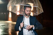 © Volodymyr - A business man checks his watch under an umbrella on a rainy night. Man in suit waits on night street holding umbrella. Business man under an umbrella, looking at watch. Man before a late meeting.