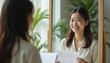© Maryna - Young asian woman practices job interview speech in front of mirror, holds paper resume. She smiles confidently, preparing for career advancement and professional growth.