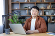 © Liubomir - Asian man smiling, recording a podcast on his laptop with a professional microphone in a comfortable home studio, creating and broadcasting audio content