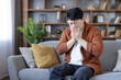 © Liubomir - Asian man sitting on a couch at home, suffering from an illness, allergy, or cold, covering his nose with a tissue while sneezing and showing discomfort