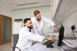 © fizkes - Digital healthcare achievement. Two young male doctors mentor and intern point at desktop monitor with smiles make diagnosis together review correct treatment result confirm positive patient progress