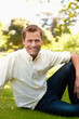 © wavebreak3 - Middle-aged man sitting on grassy lawn at park with sunlit trees, wearing light shirt and jeans
