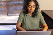 © wavebreak3 - Indian woman typing on silver laptop at office desk beside window blinds with smartphone