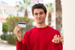 © luismolinero - Young handsome man holding a credit card at outdoors making doubts gesture while lifting the shoulders