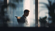 © Broccolini - A tired office worker sitting alone at a desk with a slouched posture, silhouetted against a bright window, expressing exhaustion, burnout, or mental fatigue.