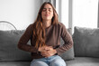 © luciano - Young woman with stomach pain sitting on the couch alone at home, holding her stomach, touching and massaging her abdomen. She has an unsightly, sickly face, and feels uncomfortable and has diarrhea.