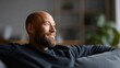 © nanskyblack - Happy bald man with beard sitting comfortably on sofa in living room, content expression, quiet relaxation and self-care at home