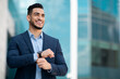 © Prostock-studio - Successful arabic entrepreneur young handsome man in suit with expensive watch on his wrist looking at copy space and smiling, walking on the street next to business building, panorama