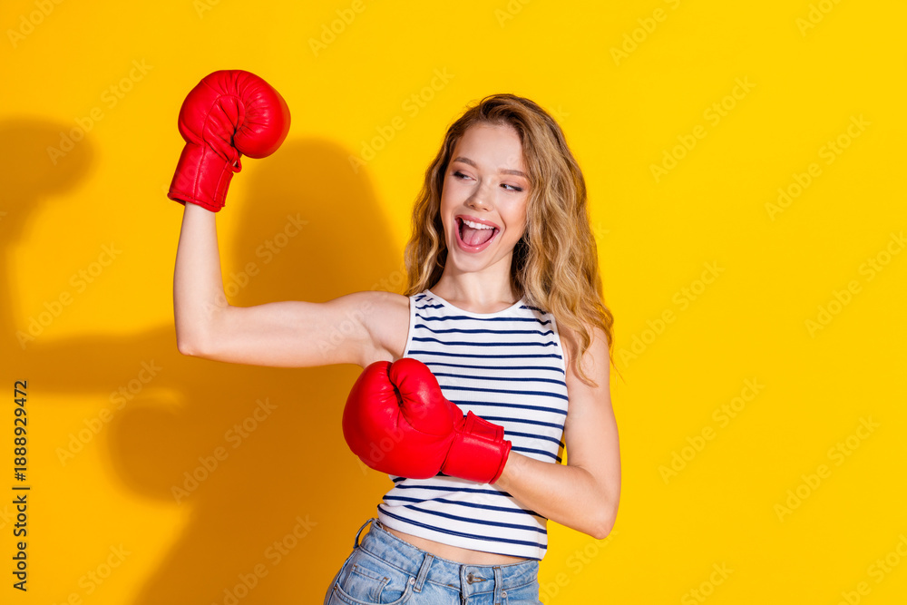 Happy young woman with boxing gloves flexing arm in a cheerful pose on a vibrant yellow background