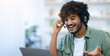 © Prostock-studio - A young man with curly hair is wearing a headset while engaging with someone on a laptop. He smiles and appears focused on his conversation. The setting has bright light and shelves in the background.