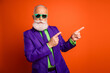 © deagreez - Vibrant retro dressed man in purple suit green shirt and sunglass points against bright orange background for fashion lifestyle promotion