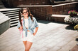 © deagreez - Young girl in casual summer outfit walks through a sunny outdoor city space with a pink backpack smiling for a fashion style portrait near stairs and brick plaza
