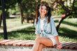 © deagreez - Young woman sits on a park bench smiling in a casual blue shirt tied at the waist with white shorts enjoying a sunny outdoor day in the park with friends