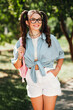© deagreez - Young stylish girl with glasses outdoors in park enjoying sunny day wearing casual blue shirt tied at waist white shorts and pink backpack