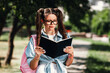 © deagreez - Young woman reads a book outdoors on a sunny day while walking through a park with a backpack and stylish glasses enjoying study and leisure in casual fashion