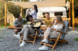 © Prostock-studio - Young multiracial friends playing guitar and singing, holding pet dog, sitting in lounge chairs near RV, camping together, having fun autumn vacation, spending time in countryside