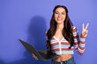 © deagreez - Young female student with glasses holds a laptop and smiles while show peace sign purple background in a casual stylish fashion shoot for lifestyle and advertising use