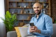 © Liubomir - Smiling man connecting online, sitting comfortably on a sofa at home, using mobile phone for communication, browsing social media, or enjoying digital content with happiness