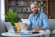 © Liubomir - Man smiling and calculating bills with a calculator, laptop, and papers while sitting on a couch, managing personal budget and household expenses in a comfortable home office setting