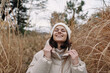 © SHOTPRIME STUDIO - Woman in a warm beige coat and knit hat stands among tall grasses, smiling under a cloudy sky, capturing a cozy outdoor moment in late autumn or early winter soft breeze.