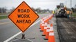 © Mariya Sorvacheva - Road work ahead sign stands prominently on roadside, surrounded by orange traffic cones, indicating construction activity and alerting drivers to upcoming road maintenance