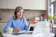 © Liubomir - Senior woman wearing a headset and looking at a laptop, gesturing while participating in an online video conference from her home kitchen, taking notes on a notebook