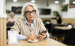 © JackF - European elderly woman drinking fresh coffee at table in light cafe and looking at mobile phone
