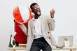 © SHOTPRIME STUDIO - Excited cheerful young African-American man in smart casual clothing celebrating with a large beach ball in modern office environment, holding fist up in triumph, smiling, positive mood, lifestyle