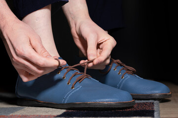  Close-up of a person's hands tying the brown shoelaces of a pair of stylish blue leather shoes. The person is sitting, preparing to go out, highlighting daily routine and self-care.