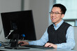 © fotolite - A smiling young Asian professional works at a desktop computer in a office environment, ideal for workplace diversity, entry level employment, corporate training, or young professional concepts.