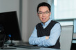 © fotolite - A confident young Asian businessman sits with arms crossed at his computer  in an office, ideal for workplace confidence, professional development, corporate culture, or career success concepts.