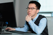© fotolite - A young Asian businessman thinks about a problem while working at his computer, ideal for problem solving, critical thinking, workplace challenges, or professional decision making concepts.