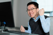 © fotolite - A smiling young Asian businessman celebrates with a fist pump gesture at his computer, ideal for workplace success, goal achievement, career milestone, or professional victory concepts.