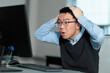© fotolite - A shocked young Asian businessman holds both hands to his head while reacting to something on his computer screen, ideal for workplace error, unexpected news, or shocking discovery concepts.