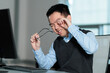 © fotolite - A fatigued young Asian man rubs his eyes while holding his glasses at his computer in the office, ideal for eye strain, workplace fatigue, digital eye fatigue, or long work hours concepts.