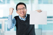 © fotolite - Young Asian man celebrates with fist pump while holding a blank sign in an office, ideal for success announcements, achievement messaging, or victory celebration concepts.