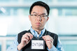 © fotolite - Young Asian intern holds up a name tag with unhappy expression, ideal for workplace dissatisfaction, internship challenges, or negative workplace experience concepts.