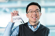 © fotolite - A young Asian man proudly holds up a lanyard with an intern badge, ideal for internship programs, new hire announcements, or corporate training concepts.