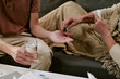 © AnnaStills - Caucasian young adult man holding glass of water while senior Caucasian woman placing pill in his palm, both sitting on sofa, hands and partial bodies visible, caregiving scene