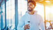 © sommersby - Smiling man in light hoodie holds water bottle while standing in bright, modern gym with large windows and sunlight streaming in, embodying fitness and well-being