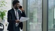 © Yuliia - African American ethnic business man angry arguing with mobile phone call problem holding papers in office furious businessman male yelling with smartphone throwing documents away looking out window