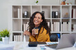 © Tj - Young woman in a yellow sweater laughing at her desk with coffee, laptop and documents, portraying productive, relaxed remote work and a confident, positive professional mood