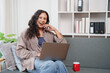 © Tj - Woman feeling happy and excited while working from home, smiling and laughing on a gray couch with a laptop and a red mug, creating a comfortable and productive remote workspace