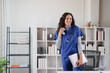 © Tj - Young happy female medical professional or nurse wearing blue scrubs and stethoscope, standing in a bright modern office while holding a laptop and smiling