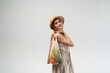 © Volodymyr_sh - Young woman with reusable mesh grocery bag over shoulder wearing straw hat and summer dress, sustainable lifestyle concept