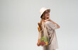 © Volodymyr_sh - Young woman in bucket hat carrying reusable mesh grocery bag with bread and vegetables, sustainable lifestyle portrait in studio