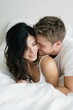 © MNStudio - Couple cuddling in bed, faces close, white sheets and soft daylight. Minimalist and bright scene focused on intimacy.
